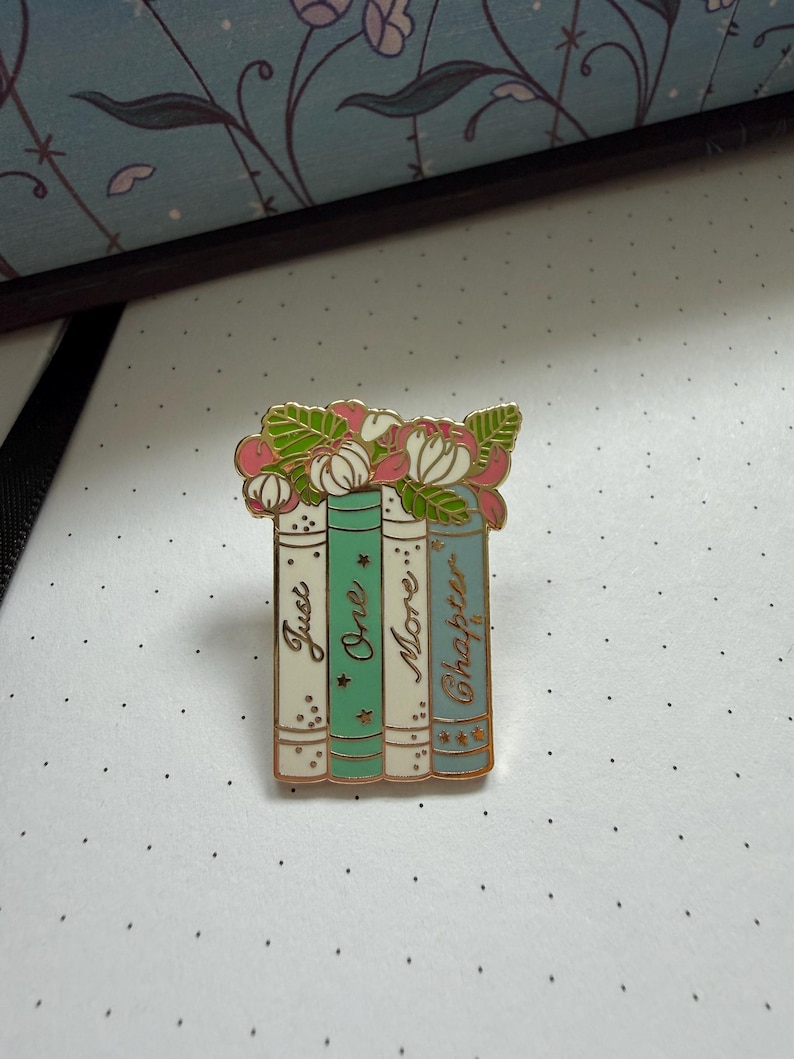 Pode incluir: Um broche de esmalte em tom dourado com quatro livros verticais com lombadas em tons pastel creme, verde menta e azul claro. Os livros s&atilde;o coroados com flores rosas e brancas e folhas verdes. O texto Just One More Chapter est&aacute; em dourado.