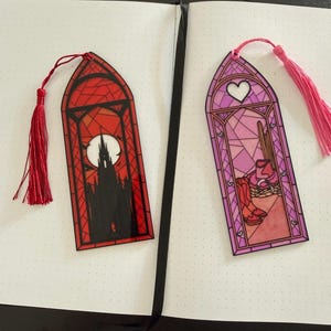 May include: Two arched bookmarks with stained glass designs. The left bookmark is red with a black silhouette of a gothic castle and a full moon. The right bookmark is pink with a heart, cowboy boots, and a hat. Both have matching tassels.