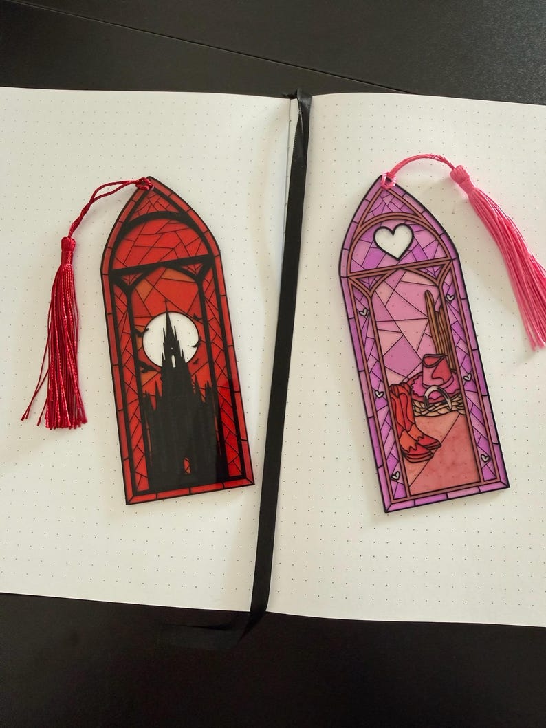 May include: Two arched bookmarks with stained glass designs. The left bookmark is red with a black silhouette of a gothic castle and a full moon. The right bookmark is pink with a heart, cowboy boots, and a hat. Both have matching tassels.