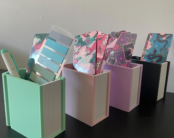 Könnte beinhalten: Vier rechteckige Schreibtisch-Organizer in Minzgrün, Hellrosa, Lavendel und Schwarz. Jeder Behälter verfügt über eine weiße, gerippte Frontplatte und enthält verschiedene Gegenstände, darunter einen hellgrünen Stift, eine blaue Farbkarte und illustrierte Karten.