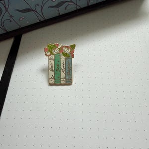 Pode incluir: Um broche de esmalte e metal dourado em forma de pilha de quatro livros, encimado por flores cor-de-rosa e brancas com folhas verdes. As lombadas dos livros s&atilde;o em tons de azul-petr&oacute;leo e creme, com o texto Just One More Chapter.