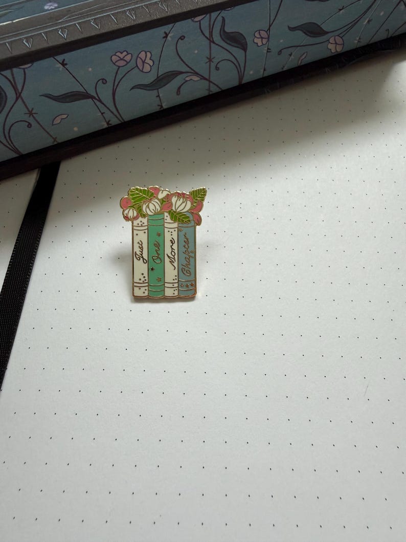 Pode incluir: Um broche de esmalte e metal dourado em forma de pilha de quatro livros, encimado por flores cor-de-rosa e brancas com folhas verdes. As lombadas dos livros s&atilde;o em tons de azul-petr&oacute;leo e creme, com o texto Just One More Chapter.