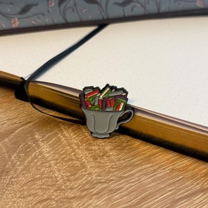 Pode incluir: Um broche de esmalte metálico em forma de xícara de chá cinza-claro preenchida com uma pilha de livros em miniatura em tons de vermelho, verde e branco. O broche apresenta um contorno preto e está posicionado na página de um caderno com uma fita preta.