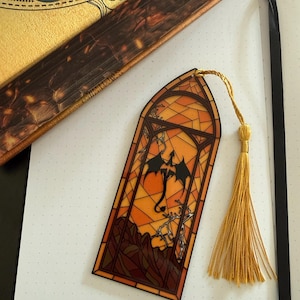 May include: A rectangular bookmark featuring a stained-glass style design with an arched top. The artwork depicts a black dragon silhouette against an orange and yellow background with dark brown mountain outlines and a gold-colored tassel attached.