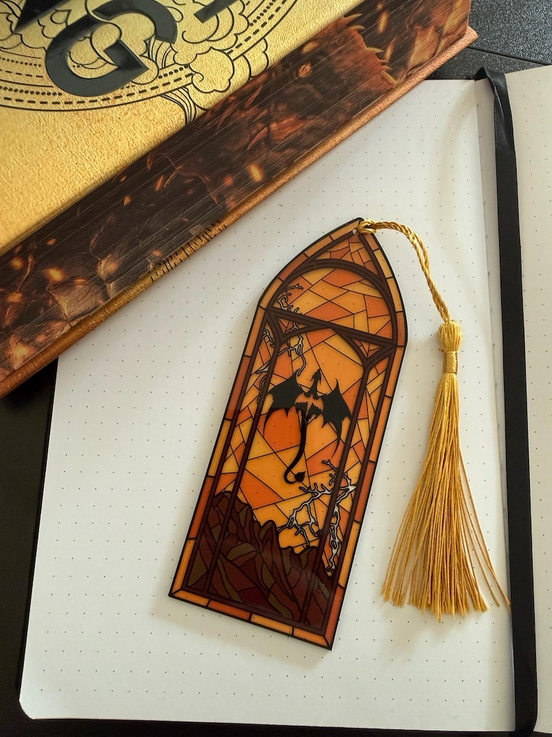 May include: A rectangular bookmark featuring a stained-glass style design with an arched top. The artwork depicts a black dragon silhouette against an orange and yellow background with dark brown mountain outlines and a gold-colored tassel attached.