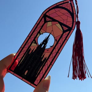 May include: A translucent bookmark shaped like a pointed arch window, featuring a black silhouette of a gothic cathedral against a large moon and a stained glass pattern in shades of pink, orange, and yellow, finished with a red tassel.