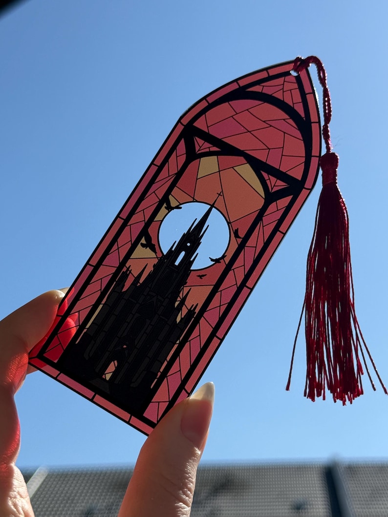 May include: A translucent bookmark shaped like a pointed arch window, featuring a black silhouette of a gothic cathedral against a large moon and a stained glass pattern in shades of pink, orange, and yellow, finished with a red tassel.