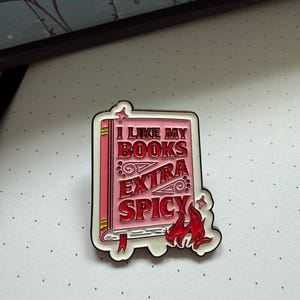 Könnte beinhalten: Eine rechteckige Emaille-Anstecknadel in Buchform mit rosa Einband und rotem Text, der I LIKE MY BOOKS EXTRA SPICY lautet. Das Design zeigt eine rote Flammengrafik in der unteren rechten Ecke, kleine Sternakzente und eine weiße Umrandung.