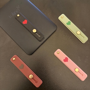 Op de afbeelding: Vier telefoonstandaards in verschillende kleuren: zwart, groen, bordeauxrood en roze. Elke standaard heeft een hartontwerp en een goudkleurige knop. Eén standaard is bevestigd aan een zwarte tablethoes met de merknaam "toling".