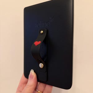 Könnte beinhalten: Schwarzer E-Reader mit strukturierter Rückseite und schwarzem Griff mit rotem Herzdesign. Auf dem E-Reader ist das Wort "tolino" aufgedruckt. Der Griff ist mit goldfarbener Hardware befestigt.