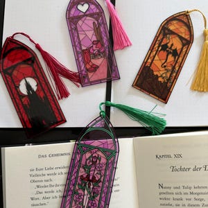 May include: Four arched bookmarks with stained glass designs and colored tassels. One features a red castle, one a pink room with boots, one an orange dragon, and one a purple floral sword design. Each bookmark has a pointed top and a hanging tassel.