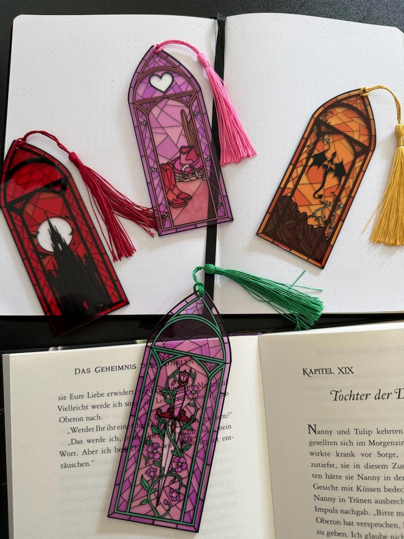 May include: Four arched bookmarks with stained glass designs and colored tassels. One features a red castle, one a pink room with boots, one an orange dragon, and one a purple floral sword design. Each bookmark has a pointed top and a hanging tassel.