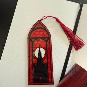May include: A rectangular bookmark with a pointed arch top, featuring a stained glass style design in red and black. The image depicts a dark gothic castle silhouette against a large white full moon, finished with a red thread tassel at the top.