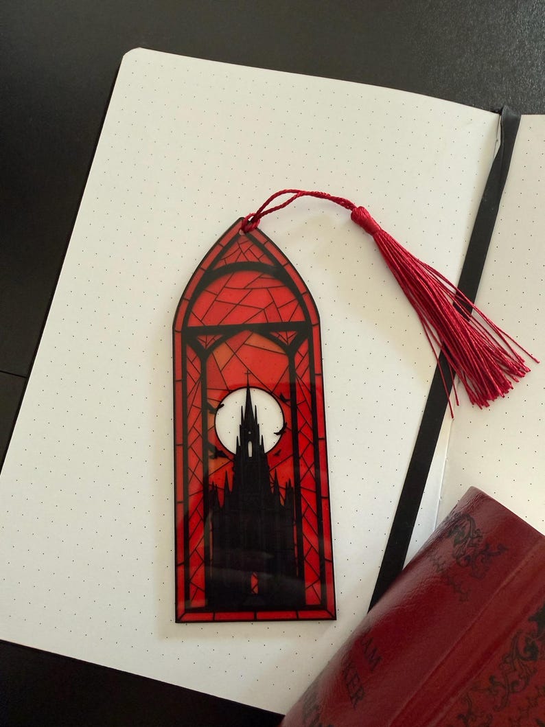 May include: A rectangular bookmark with a pointed arch top, featuring a stained glass style design in red and black. The image depicts a dark gothic castle silhouette against a large white full moon, finished with a red thread tassel at the top.