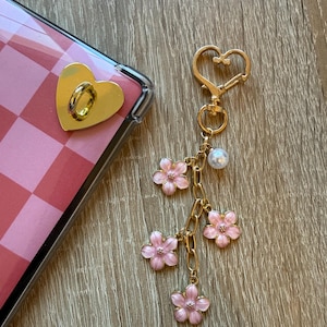 Puede incluir: Un soporte de teléfono dorado en forma de corazón y un llavero dorado con dijes de flores rosas y una perla. La funda del teléfono tiene un patrón de cuadros rosas y blancos.