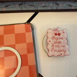 Puede incluir: Un soporte para teléfono rectangular con borde festoneado y un patrón de cuadros rosa claro. El diseño incluye una ilustración de cerezas rojas con un lazo y el texto "Buy me books & tell me I'm pretty" en letra cursiva roja con corazones.