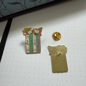 Pode incluir: Um broche de esmalte em tom dourado com tr&ecirc;s livros empilhados e flores cor-de-rosa no topo. As lombadas s&atilde;o verde-claro, azul-petr&oacute;leo e branco, com o texto "Just One More Chapter" escrito. Um fecho dourado separado e o verso s&atilde;o vis&iacute;veis.