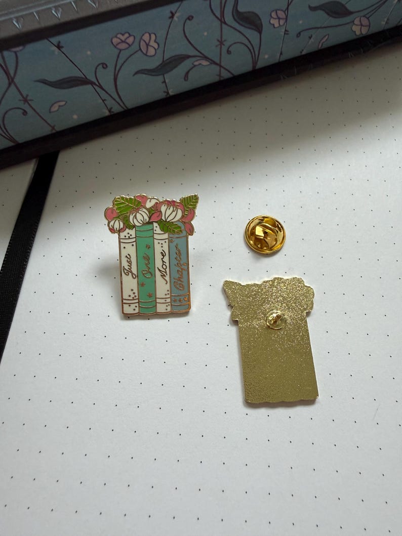 Pode incluir: Um broche de esmalte em tom dourado com tr&ecirc;s livros empilhados e flores cor-de-rosa no topo. As lombadas s&atilde;o verde-claro, azul-petr&oacute;leo e branco, com o texto "Just One More Chapter" escrito. Um fecho dourado separado e o verso s&atilde;o vis&iacute;veis.