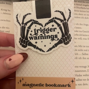 Könnte beinhalten: Ein schwarzer und weißer magnetischer Lesezeichen mit einem Skelett-Hand-Design. Das Lesezeichen zeigt den Text "trigger warnings" in einem Herz.
