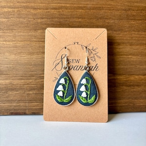 May include: A pair of teardrop-shaped earrings featuring embroidered lily of the valley designs. The earrings have a dark blue background with white flowers and green leaves, set in silver-colored frames. The earrings are displayed on a brown card with the text "Sew Savannah".