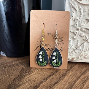 May include: Embroidered teardrop earrings featuring a lily of the valley design. The earrings have a dark blue background with white and green floral embroidery, set in a silver and gold frame. The earrings are displayed on a brown card.