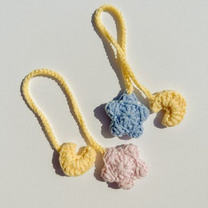 Pode incluir: Dois enfeites de croch&ecirc; feitos &agrave; m&atilde;o. Um &eacute; uma estrela azul clara e o outro uma flor rosa. Ambos est&atilde;o presos a um la&ccedil;o amarelo. Um enfeite amarelo em forma de cora&ccedil;&atilde;o tamb&eacute;m &eacute; vis&iacute;vel. Os enfeites est&atilde;o em um fundo branco.