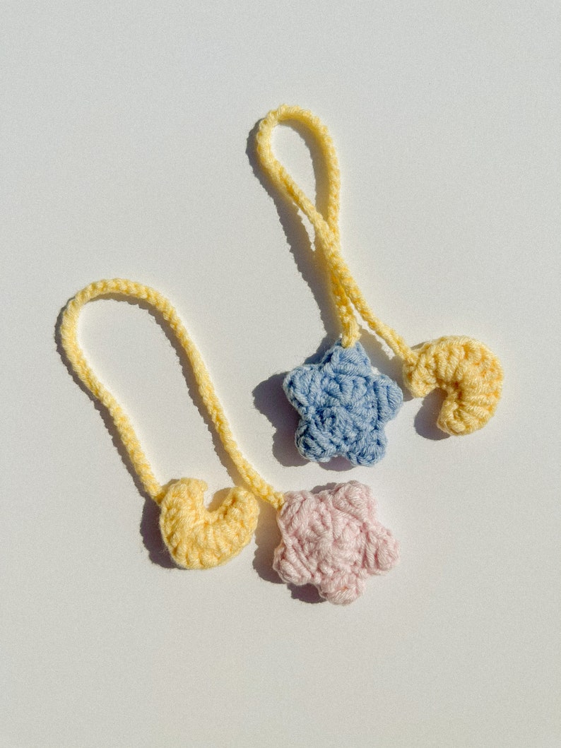 Pode incluir: Dois enfeites de croch&ecirc; feitos &agrave; m&atilde;o. Um &eacute; uma estrela azul clara e o outro uma flor rosa. Ambos est&atilde;o presos a um la&ccedil;o amarelo. Um enfeite amarelo em forma de cora&ccedil;&atilde;o tamb&eacute;m &eacute; vis&iacute;vel. Os enfeites est&atilde;o em um fundo branco.