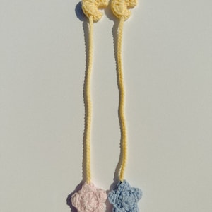 Pode incluir: Dois marcadores de croch&ecirc; feitos &agrave; m&atilde;o. Cada marcador apresenta uma forma de lua crescente amarela conectada a um cord&atilde;o amarelo, que leva a uma estrela. Uma estrela &eacute; rosa e a outra azul. Os marcadores s&atilde;o exibidos em uma superf&iacute;cie branca.