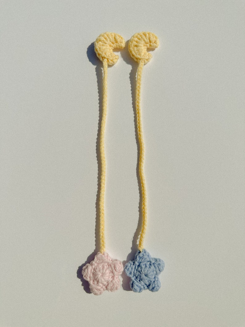 Pode incluir: Dois marcadores de croch&ecirc; feitos &agrave; m&atilde;o. Cada marcador apresenta uma forma de lua crescente amarela conectada a um cord&atilde;o amarelo, que leva a uma estrela. Uma estrela &eacute; rosa e a outra azul. Os marcadores s&atilde;o exibidos em uma superf&iacute;cie branca.