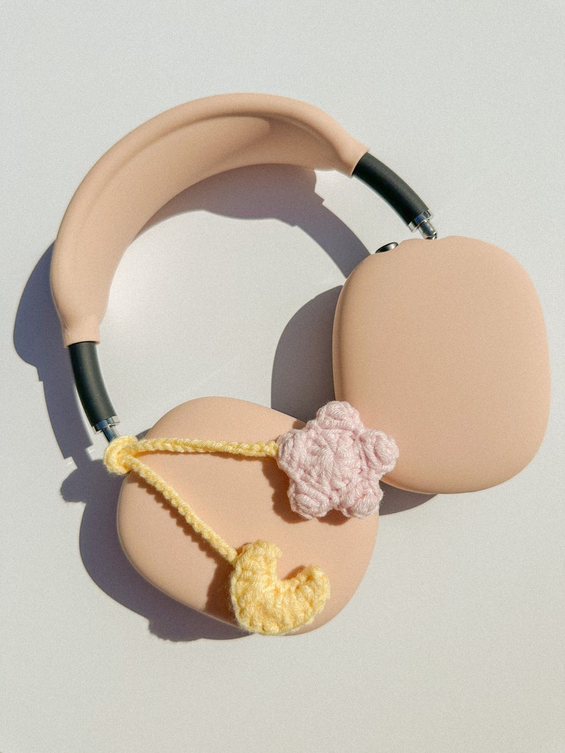 Pode incluir: Fones de ouvido circum-aurais cor de rosa claro com faixa de cabe&ccedil;a e conchas pretas. Os fones de ouvido s&atilde;o decorados com um cora&ccedil;&atilde;o de croch&ecirc; amarelo e uma estrela de croch&ecirc; rosa, conectados por uma corrente de croch&ecirc; amarela.