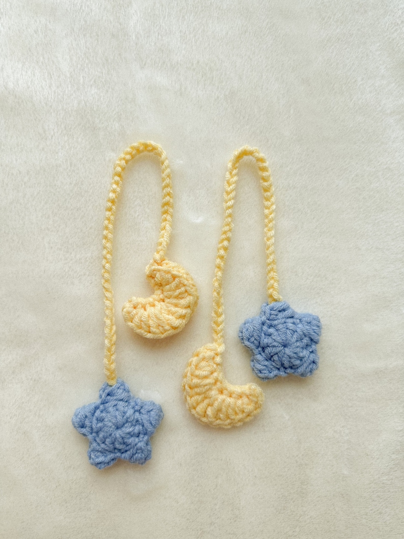 Pode incluir: Dois enfeites de croch&ecirc; feitos &agrave; m&atilde;o com um tema celestial. Um tem uma estrela azul clara e uma lua crescente amarela. O outro tem uma lua crescente amarela e uma estrela azul clara. Ambos t&ecirc;m um cord&atilde;o amarelo para pendurar.