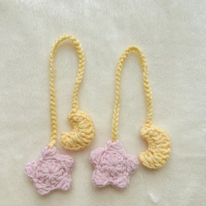 Pode incluir: Dois ornamentos de croch&ecirc; feitos &agrave; m&atilde;o. Cada ornamento apresenta uma estrela rosa e uma lua crescente amarela, conectadas por uma corrente amarela. Os ornamentos s&atilde;o exibidos em uma superf&iacute;cie branca e macia.
