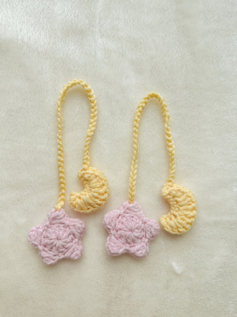Pode incluir: Dois ornamentos de croch&ecirc; feitos &agrave; m&atilde;o. Cada ornamento apresenta uma estrela rosa e uma lua crescente amarela, conectadas por uma corrente amarela. Os ornamentos s&atilde;o exibidos em uma superf&iacute;cie branca e macia.