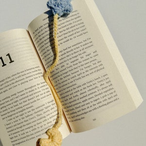 Pode incluir: Um marcador de livros de croch&ecirc; feito &agrave; m&atilde;o com uma estrela azul clara e uma base amarela arredondada. O marcador de livros est&aacute; colocado em um livro aberto, com os n&uacute;meros das p&aacute;ginas vis&iacute;veis. O livro est&aacute; aberto na p&aacute;gina 151.