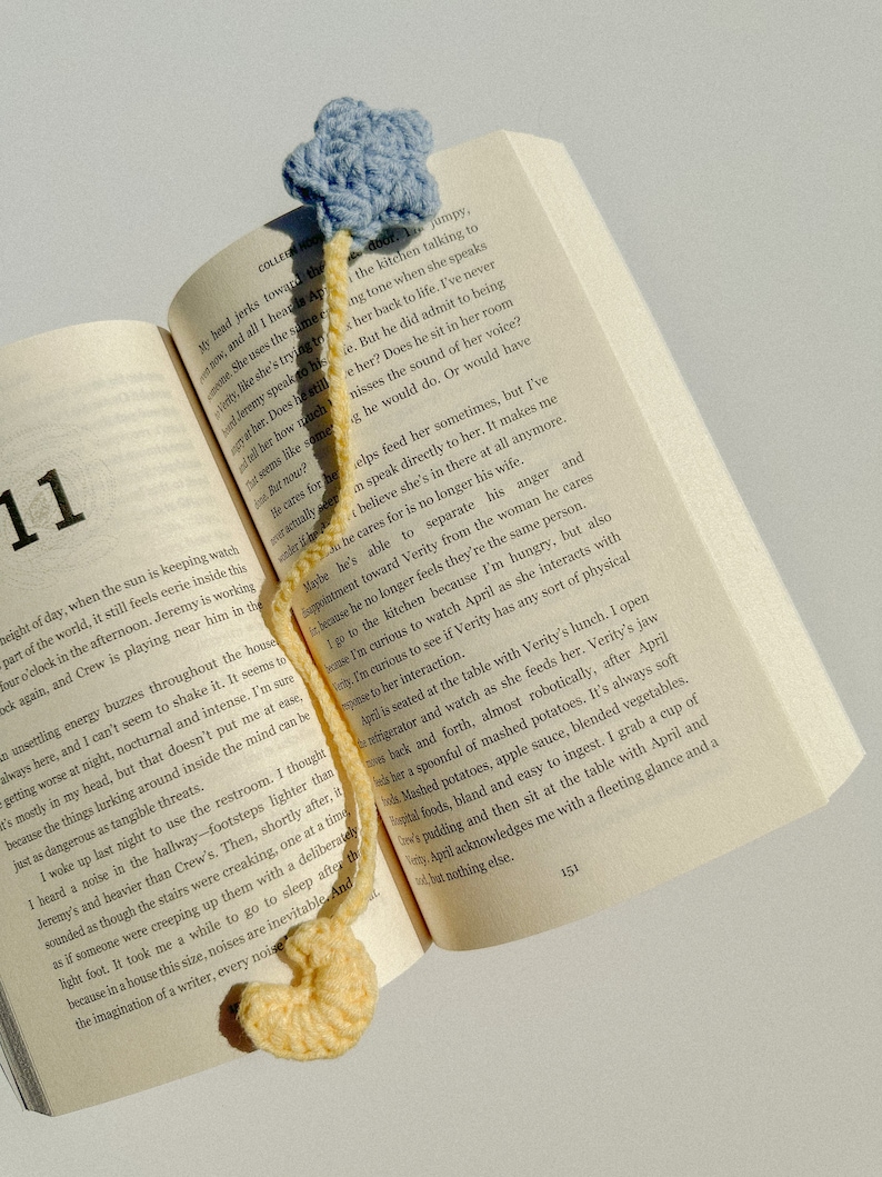 Pode incluir: Um marcador de livros de croch&ecirc; feito &agrave; m&atilde;o com uma estrela azul clara e uma base amarela arredondada. O marcador de livros est&aacute; colocado em um livro aberto, com os n&uacute;meros das p&aacute;ginas vis&iacute;veis. O livro est&aacute; aberto na p&aacute;gina 151.
