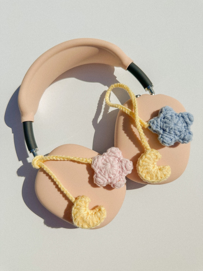 Pode incluir: Fones de ouvido rosa claro com detalhes em preto e pingentes de croch&ecirc;. Os pingentes incluem uma lua crescente amarela, uma estrela rosa e uma estrela azul. Os fones de ouvido est&atilde;o em uma superf&iacute;cie branca.