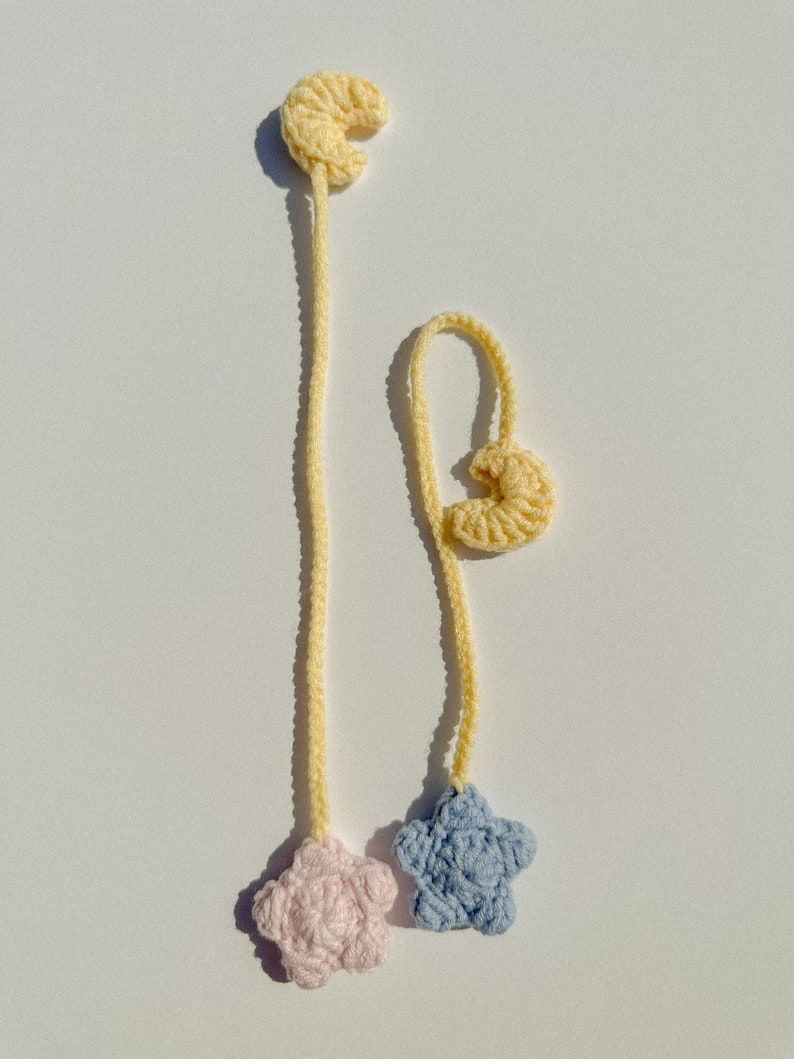 Pode incluir: Dois marcadores de croch&ecirc; feitos &agrave; m&atilde;o. Cada marcador apresenta uma forma de crescente amarelo no topo, conectado a um cord&atilde;o amarelo. Um cord&atilde;o termina com uma estrela rosa, o outro com uma estrela azul. O fundo &eacute; branco.