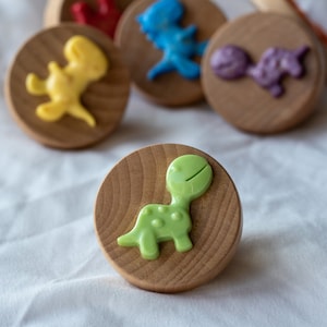 Puede incluir: Cinco sellos de juguete de madera con diferentes diseños de dinosaurios. Los sellos están pintados en varios colores, incluyendo amarillo, azul, rojo, morado y verde. Los sellos están dispuestos en una fila sobre una superficie blanca.
