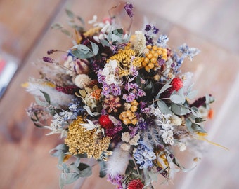 May include: A bouquet of dried flowers in shades of purple, yellow, and red. The bouquet includes dried grasses, lavender, and other wildflowers.