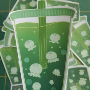 May include: A green plastic cup with a straw filled with a green liquid and white jellyfish. The cup has a white rim and a white lid.