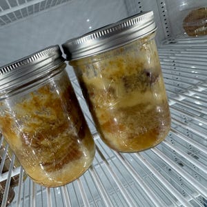 Könnte beinhalten: Zwei Glasbehälter mit silbernen Deckeln, gefüllt mit einem gelben und braunen, geschichteten Dessert, im Kühlschrank aufbewahrt.
