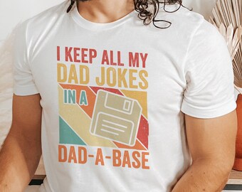 May include: White t-shirt with the text "I KEEP ALL MY DAD JOKES IN A DAD-A-BASE" in orange, red, and yellow. The graphic includes a retro-style floppy disk design. The shirt is worn by a person.