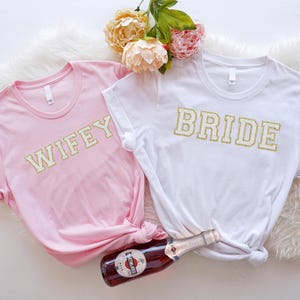 May include: Two white t-shirts with gold glitter lettering. One shirt says "Bride" and the other says "Wifey".