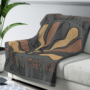 May include: A gray throw blanket draped over a white sofa. The blanket features a retro design with the text "Fill the Air" and a stylized graphic in shades of brown, tan, and gray. A large potted plant is visible in the background.