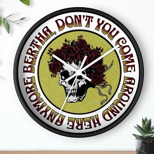 May include: A black-framed wall clock featuring a skull adorned with a crown of red roses. The clock face has the text "MORE BERTHA, DON'T YOU COME AROUND HERE ANYMORE". The background is a light green circle.