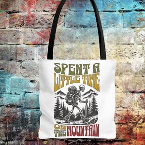 May include: White tote bag with a black handle featuring a skeleton hiker design. The design includes the text "Spent a Little Time on the Mountain" in a retro font, with a mountain and tree illustration.