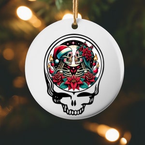 May include: White ceramic Christmas ornament featuring a stylized skull design. Inside the skull is a couple of skeletons with festive holiday decorations, including a Santa hat, flowers, and candles. The ornament hangs from a brown string.