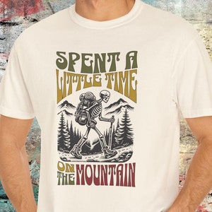 May include: A white t-shirt with a graphic of a skeleton hiking in the mountains. The text "Spent a little time on the mountain" is printed in a retro font.
