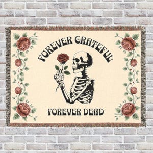 May include: Woven throw blanket with a cream background featuring a skeleton holding a red rose. The words "FOREVER GRATEFUL" arch above the skeleton, and "FOREVER DEAD" is below. Floral accents and fringe complete the design.