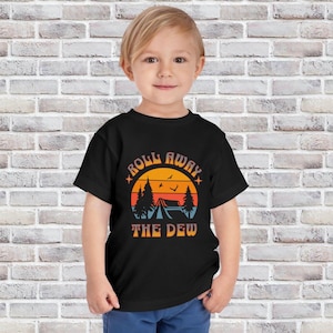May include: A black toddler t-shirt featuring a camping-themed graphic. The design includes a sunset, trees, a tent, and the text "Roll Away The Dew" in orange and blue. The shirt is worn by a young child.
